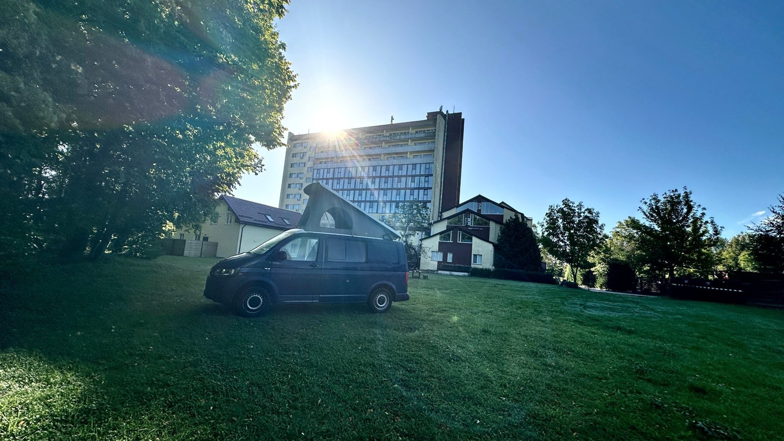Dunkler VW-Bulli mit Aufstelldach auf einer Wiese vor einem Hotelhochhaus in Kaliningrad, Gegenlicht